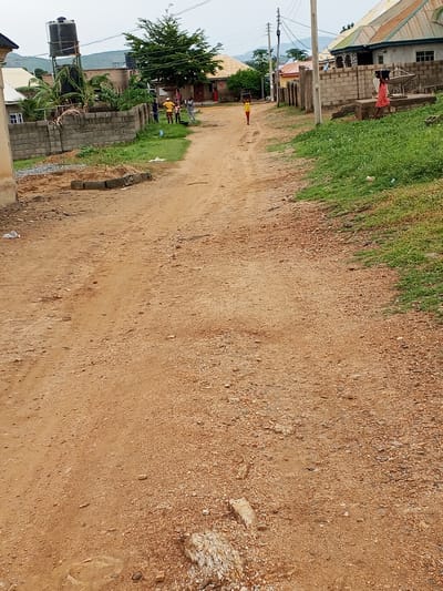 The image depicts a dirt road in Ashata, Nigeria. On the left side of the road, a tall water tank on a metal structure is visible, along with a brick wall and some greenery. Several individuals, including men and children, are gathered near the wall. The road itself is unpaved and appears to be a common thoroughfare in the area, with scattered stones and debris. In the distance, more buildings and a lush green tree can be seen, suggesting a residential or semi-rural setting. The sky is overcast. Further down the road, a young person in a pink dress is walking, carrying an object on their head, and another individual in a yellow shirt is also visible in the background. The overall scene conveys a sense of everyday life in the depicted location.
