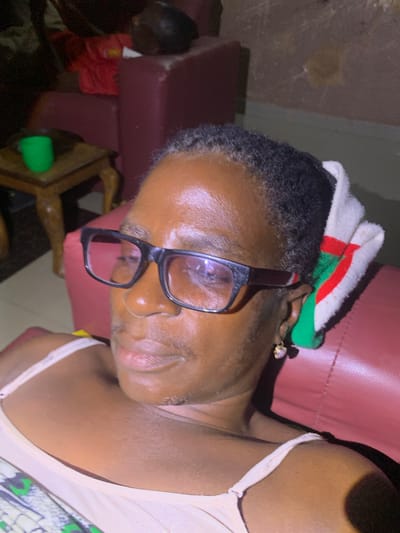A close-up, dimly lit, indoor, eye-level photograph captures a Black woman wearing glasses and a headscarf. She is lying down, her head resting on a red cushioned surface, likely a sofa or chair. The woman is facing the camera, with her right side more prominent. She has short, dark, curly hair, and is wearing black-rimmed glasses with thick frames. A white headscarf with red and green stripes is wrapped around the back of her head, with a portion extending over her right ear. A small, gold earring with a pearl-like stud is visible on her right earlobe. Her skin appears oily or sweaty, reflecting the light. She is wearing a light-colored tank top with thin straps. Her mouth is slightly open, and her expression is neutral. In the background, to the left, a small wooden table with carved legs holds a bright green cup. Behind the table, a red fabric item is visible, possibly clothing. The overall setting suggests a residential interior in Barakin Sabawa, Nigeria. The lighting is artificial and casts shadows, contributing to the overall low-light conditions of the image.