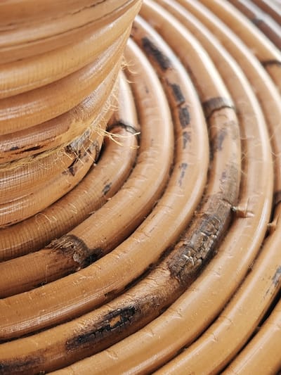 The image is a close-up, abstract shot of a woven rattan object. The texture of the rattan is the dominant feature, showcasing its natural fibers and a glossy finish. Some areas of the rattan appear darker and more weathered, with visible cracks and frayed strands, suggesting age or wear. The object is cylindrical, with the weaving forming concentric, curved lines that recede into the background. The lighting highlights the curved surfaces and creates subtle shadows. The image does not contain any identifiable elements that would allow for a specific location to be determined, nor does it depict any people, actions, or interactions.