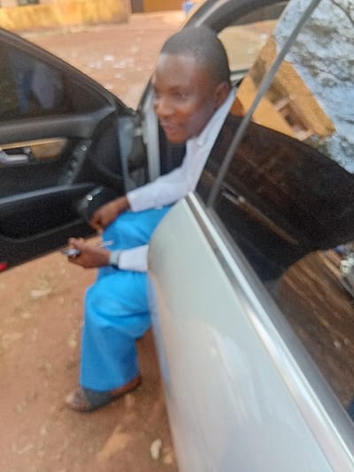 The image shows a person leaning into the open front passenger door of a light-colored car. The image is oriented vertically, rotated 90 degrees counter-clockwise.
The central figure is a young person, possibly male, with dark skin and short, dark hair. They are dressed in a white long-sleeved shirt and bright blue trousers or a uniform. Their upper body is angled downwards and partially obscured by the car's interior as they lean into the vehicle. Their right arm is visible, bent at the elbow, and they appear to be holding a small, dark object, possibly a pen or phone, in their right hand. The person is wearing dark, open-toed shoes or sandals on their feet, which are positioned on the ground outside the car.
The car is light-colored, possibly white or silver. The front passenger door is wide open, revealing a dark interior. The exterior side panel of the car is visible.
The ground beneath the person's feet and extending into the foreground and background is a reddish-brown, dusty surface, consistent with dirt or unpaved earth. In the background, beyond the car, there are some blurry, indistinct structures. A light-colored, rough surface is visible in the upper left, and a brownish, possibly wooden or brick-like structure, appears behind the person's head. The overall lighting suggests it is daytime. No text is visible in the image.