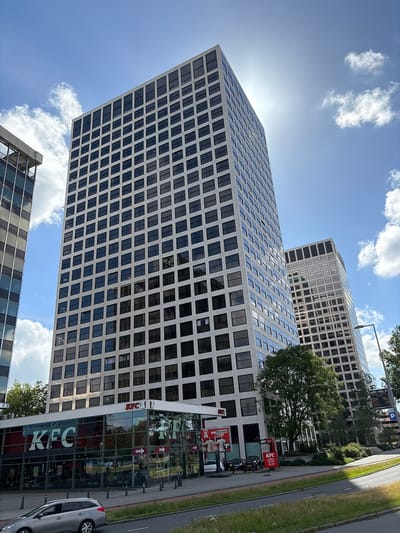 In Rotterdam, Netherlands, two tall, modern office buildings with repetitive window patterns dominate the scene under a bright, sunny sky with scattered clouds. The building on the left is mostly obscured by the building on the right, which features a grid of dark windows against a light-colored facade. In the foreground, a single-story KFC restaurant with large glass windows is situated at the base of the taller building. The KFC's branding is clearly visible, including the iconic logo and signage. A silver car is parked on the left side of the street in front of the KFC. The environment includes a paved road, a grassy median, and some trees and bushes surrounding the buildings. A streetlamp is visible on the right side of the image. The overall impression is an urban commercial area on a clear day.
