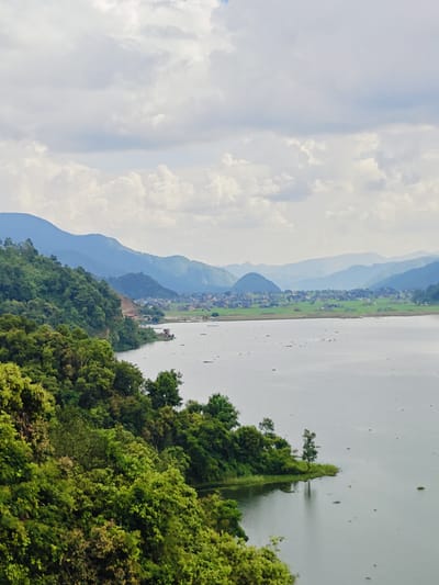 The image shows a panoramic view of a serene lake, likely Phewa Lake, in Rupakot, Nepal, under a cloudy sky. Lush green hills covered in dense vegetation slope down to the water's edge on the left side of the frame. In the distance, rolling mountains form a hazy blue backdrop, with a small town nestled between the hills and the lake. The water surface of the lake is calm, with a few small, dark objects, possibly boats or buoys, scattered across it. The foreground is dominated by the vibrant greenery of the trees and foliage. The overall impression is one of natural beauty and tranquility.