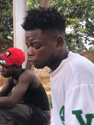 This image shows two males outdoors in Oraukwu, Nigeria. In the foreground, a young adult male with dark skin, short, faded black curly hair, and light facial stubble is depicted. He wears a white, textured long-sleeved shirt with green letter appliques, one appearing as a 'V'. His gaze is directed downward, with eyes seemingly closed or cast down, and visible forehead creases.
To his left and slightly behind him sits a younger male with dark skin, wearing a red baseball cap with a gold and black emblem, a black tank top, and dark pants. He faces left, away from the camera.
The environment consists of a reddish-brown dirt ground. Behind the seated male, a metal fence is visible, while a light-colored vertical structure stands behind the primary subject. Further back, green foliage from trees and a reddish-brown wall are present under bright daylight.