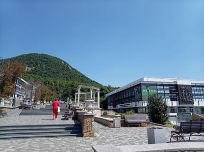 In Zheleznovodsk, Russia, a woman in a red outfit walks up a wide, paved stone staircase. The staircase leads to a park-like area with stone benches and decorative planters. A large, modern building with a glass facade stands to the right of the stairs, featuring several posters and a large metal relief sculpture on its exterior. In the background, a lush green mountain slopes upwards under a clear blue sky. The scene appears to be a public space, possibly a promenade or a park leading to a cultural center.