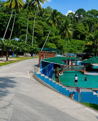 This is a medium shot, eye-level, full-color photograph of a swimming pool area in Silaiyajae, Indonesia. The swimming pool is a bright turquoise color and has a wooden deck surrounding parts of it. On the deck, two young boys are visible. One boy, wearing a white tank top and black shorts, stands at the edge of the deck. Another boy is in the water. A woman wearing a headscarf and a floral dress is standing on blue steps leading down to the pool, looking out at the water. To the right of the pool, there is a covered pavilion with a green roof, and in the background, there are lush green trees and palm trees under a clear blue sky with scattered clouds. A paved walkway curves from the left foreground towards the pool area. Several blue umbrellas are also visible in the distance, as well as a building with the words "MUSHOLLA" on it. A wooden fence with horizontal planks separates the walkway from a grassy area.