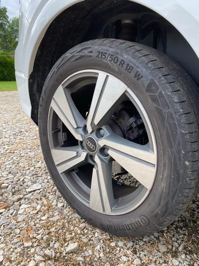 This image captures a close-up of the front wheel and tire of a white Audi vehicle, parked on a gravel surface. The scene is outdoors, brightly lit, suggesting daytime with fair weather. Green foliage and a hint of a tree are visible in the background, implying a setting in Courpière, France, possibly a driveway or rural road.
The primary subjects are the dark grey alloy wheel with the Audi rings logo prominently displayed on the center cap, and a black Continental tire. The tire tread pattern is visible and shows some wear and dirt. The tire also has a visible valve stem.
Notable details are the numerous markings on the tire sidewall, clearly readable from the image and its crops:
* Manufacturer: **Continental**
* Tire model: **ContiContact 6**
* Tire size specification: **215/50 R18 92W** (width 215mm, aspect ratio 50%, radial construction, 18-inch rim diameter, 92 load index, W speed rating).
* Tire performance ratings: **TREADWEAR 420 TRACTION A TEMPERATURE A**
* Maximum inflation pressure: **MAX INFLATION PRESSURE 350 KPA (51 PSI)**
* Maximum load capacity: **MAX LOAD 630 KG (1389 LB)**
* DOT code: **DOT JCP 0829JL 0824**, indicating the tire was manufactured in the 8th week of 2024.
* The word **"OUTSIDE"** is also visible, indicating the correct mounting direction.
No activity is taking place; the car appears to be stationary. The focus is purely on the technical details of the tire and wheel.