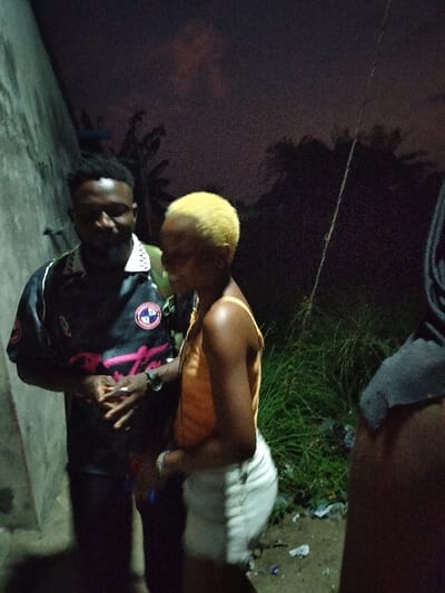 At dusk, a man and a woman stand outdoors. The man is on the left wearing a dark shirt with a design. The woman is next to him, wearing a light-colored tank top and white shorts, with blonde hair. Another individual is partially visible on the right side. Behind them are trees, bushes, and a dark sky with some clouds. The photo appears to have been taken in dim lighting.