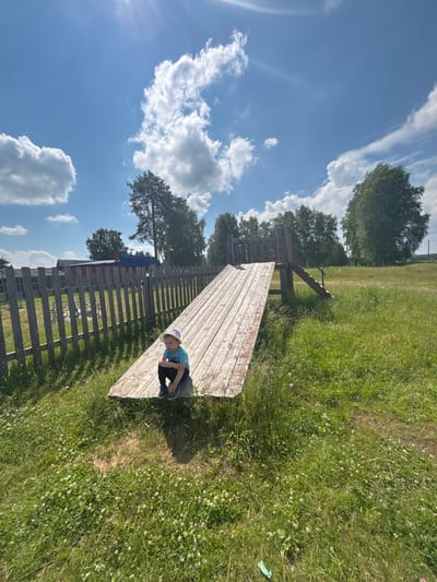 A young boy wearing a light blue t-shirt, dark pants, a light-colored hat, and dark shoes sits on the end of a wooden slide in a grassy outdoor area. The slide is constructed from light-colored wooden planks and has stairs leading up to it on the right side. The boy is facing away from the camera, with his knees tucked up towards his chest, and his arms wrapped around his legs. He appears to be looking downwards.
Behind the slide and to the left is a wooden fence with vertical posts and horizontal planks, and behind the fence, a red building with a blue roof is visible. The environment is rural, with a large expanse of green grass in the foreground and middle ground, dotted with small white flowers. In the background, numerous trees, both deciduous and coniferous, are present. The sky is bright blue with scattered white, fluffy clouds.
The image depicts a scene in a village or rural settlement, consistent with the landscape typically found in the outskirts of a city like Kurgan, Russia. The overall atmosphere is one of a sunny, summer day. There are no other people visible in the image. The lighting suggests it is daytime, likely late morning or early afternoon, with the sun casting shadows from the slide onto the grass.