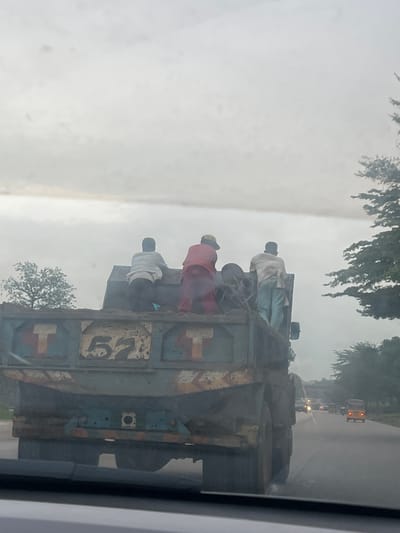 In Karmajiji, Nigeria, a dirt-filled truck with three people standing in the back drives on a road under an overcast sky. The truck's side has the number "57" painted on it, along with what appears to be the letter "T" on either side. Two of the individuals are wearing light-colored shirts and dark pants, while the third person is wearing a red garment and a yellow hat. Other vehicles, including cars and a yellow tricycle, are visible on the road ahead, suggesting moderate traffic. Trees line the side of the road, and the overall atmosphere appears to be hazy or dusty.