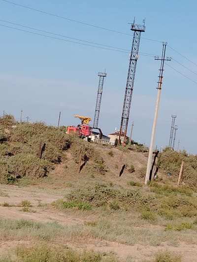 The image shows a construction site with several tall metallic towers, some appearing to be electrical pylons and others possibly part of a crane or tower structure. A red crane truck is visible on a hillside covered in dry brush and vegetation. In the background, there are buildings and more electrical pylons extending into the distance. The sky is clear and blue. The scene is located in the city of Töretam, Kazakhstan. The terrain is uneven and arid, characteristic of the steppe region. There are no people clearly visible in the image. The overall impression is one of infrastructure development or maintenance in a rural or semi-rural setting.
