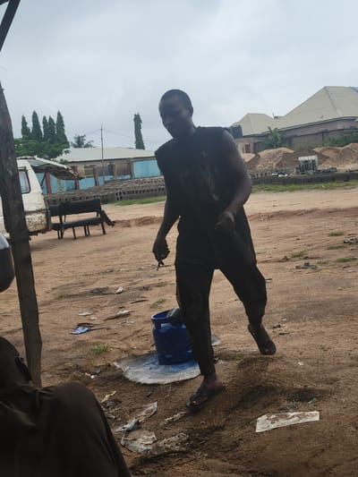 The image depicts an outdoor scene in Kwamba, Nigeria, under an overcast sky. A Black man is the central figure, dressed in dark clothing. He is shown mid-stride, with his right arm slightly bent and his hand open. He is wearing dark sandals. A blue gas cylinder is visible on the ground near his feet. The ground is composed of dirt and sand, with scattered debris including plastic wrappers and what appears to be a piece of white paper. In the background, a white van is parked on the left, and a dark bench is positioned behind it. Beyond the immediate area, there are piles of sand and building materials, along with several low-rise buildings. Trees are visible in the distance, and some construction activity is implied by the presence of building blocks. The overall impression is one of a somewhat dusty, utilitarian outdoor space.