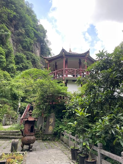 A downward angled medium shot captures a pagoda nestled in lush greenery in Wulingyuan, China. The pagoda features a red and beige construction and a multi-tiered roof. The surrounding environment is densely vegetated with trees and foliage. A stone path leads towards the pagoda, and an ornate, dark-colored incense burner stands to the right of the path. A stone tablet inscribed with golden lettering is behind the incense burner. The sky is visible, featuring a mix of clouds and blue patches.