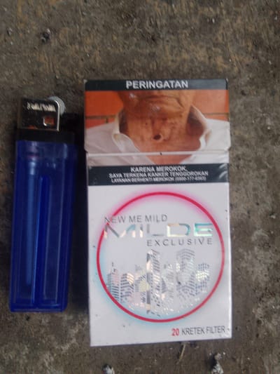 A blue gas lighter lies next to a pack of "NEW ME MILD EXCLUSIVE" cigarettes on a textured gray surface, possibly concrete or asphalt. The cigarette pack features an image of a person's face on the top portion, with the words "PERINGATAN" (Warning) visible. Below the face, text reads "KARENA MEROKOK, SAYA TERKENA KANKER TENGGOROKAN, LAWAN BERHENTI MEROKOK (0800-177-6565)" (Because of smoking, I have throat cancer, Fight smoking). The brand name "NEW ME MILD" is prominent, accompanied by "EXCLUSIVE" and an illustration of city buildings. The bottom of the pack indicates "20 KRETEK FILTER". The lighting is harsh, creating some shadows. The exact location in Cisarua, Indonesia is not discernible from the image itself.