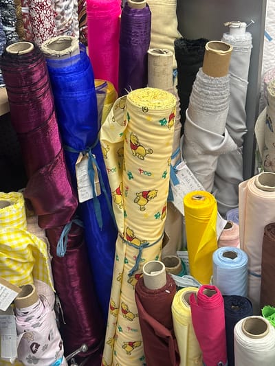 A densely packed collection of fabric rolls fills the frame. The fabrics vary in color, pattern, and texture, creating a visually rich tableau. Prominent colors include yellow, blue, purple, pink, and white. Several rolls feature patterns, such as Winnie-the-Pooh characters, floral designs, and checks. The fabric rolls are stacked haphazardly, with some labels and tags visible. The rolls appear to be in a warehouse. There is no evidence of the image being located in the city of Sevenoaks, United Kingdom.