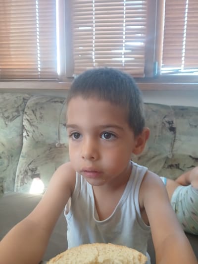 A young boy with brown hair and brown eyes is looking to the side, with his mouth slightly open. He is wearing a white tank top and is sitting on a couch with a patterned fabric. In front of him, there is a slice of bread. The background shows closed horizontal blinds on a window. The scene appears to be indoors, possibly in a home in Obrochishte, Bulgaria.