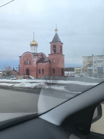 The image displays a red brick Orthodox church with an adjacent bell tower, located in Krasnyy Oktyabr', Russia, under an overcast sky. The church features a large central golden onion dome atop a white drum, capped with a golden Orthodox cross. To its right, a taller, square red brick bell tower with a dark conical roof is visible, also surmounted by a smaller golden spire and cross; multiple bells are discernable within its open arches. The church's facade exhibits arched windows and a prominent arched main entrance. Sections of the red brick show lighter, possibly unfinished or weathered, areas.

The ground surrounding the church and the foreground roadside are covered in patches of dirty snow and dark asphalt. A low fence runs along the perimeter of the church grounds. In the background to the right, a multi-story, light-colored residential apartment building with balconies stands. Further to the left and in the distant background, bare trees and other smaller structures are present. The lower right foreground is obscured by internal components of a vehicle, including a gray dashboard, an A-pillar, and a reflection of a side mirror, indicating the photograph was captured from within a car. A dark utility line extends diagonally across the upper left quadrant of the frame. No individuals, explicit actions, or interactions are visible.