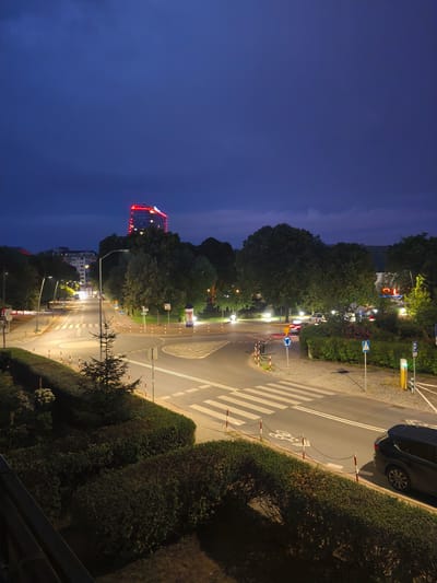 The image captures a street intersection in Szczecin, Poland, during the twilight hours. A multi-story building with red lights illuminating its top floors stands prominently in the background, characteristic of the building at al. Papieża Jana Pawła II. The street is illuminated by streetlights, casting a glow on the road, pedestrian crossings, and surrounding vegetation. Several cars are visible on the road, one dark SUV parked near a pedestrian crossing. The scene is characterized by a mix of urban infrastructure and greenery, with trees lining the sides of the street and manicured hedges in the foreground. The sky is a deep blue, indicating the transition from day to night.