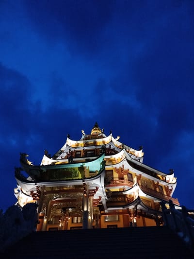 A low-angle shot depicts the illuminated Hoằng Pháp Temple in Phú An, Vietnam against a dark blue, clouded sky. The temple is multi-tiered with ornate roofs and intricate details. Lights highlight the architecture, including pillars, balconies, and decorative elements. Stairs lead up to the temple, and stone or concrete structures are visible near the base. The time of day is likely dusk or night.