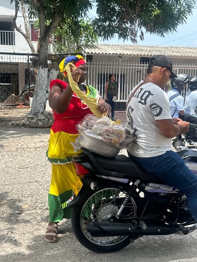 In Perímetro Urbano Barranquilla, Colombia, a woman wearing a vibrant red, yellow, and green dress and a colorful headscarf stands next to a motorcycle. She is holding a yellow cloth and a plastic-wrapped bundle of food, possibly bread or pastries, placed on the back of the motorcycle. A man wearing a white t-shirt with black lettering and a black baseball cap is seated on the motorcycle, looking forward. He is wearing blue jeans and appears to be holding a phone. Other individuals and motorcycles are visible in the background, suggesting a street scene in a populated area. The environment includes a tree with white-painted bark and a building with a white tiled roof and barred windows. The ground is paved with textured concrete.