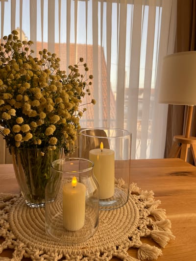This image depicts a still life arrangement on a wooden surface, likely a table, in a well-lit interior. The primary subjects are a bouquet of delicate, pale yellow flowers and two cylindrical, cream-colored candles housed within clear glass hurricanes. The flowers, abundant and bushy, are arranged in a clear glass vase that is mostly out of frame but appears to be quite large. One candle is positioned within a taller hurricane, and the other within a shorter, wider one. Both candles have a warm, artificial flame, suggesting they are LED or battery-operated.

The backdrop includes a horizontally striped sheer curtain, through which glimpses of outdoor scenery are visible, including what appears to be a tiled roof and possibly other buildings, hinting at an urban or suburban setting. The natural wood grain of the table is prominent, adding a rustic element. A macrame placemat with a fringed edge is placed beneath the candle holders, further enhancing the textural and decorative aspects of the arrangement.

The lighting suggests it could be daytime, possibly late afternoon, given the soft, diffused light filtering through the curtains. There are no people visible in the image, and the overall mood is serene and decorative, focused on creating a warm and inviting ambiance. The setting appears to be a home interior, possibly a dining room or living area. There is no visible text in the image.