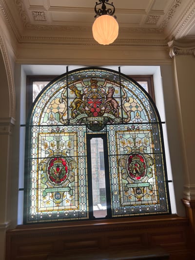 The image shows a large, ornate stained-glass window, likely in a historic building. The window is arched at the top and divided into several sections. The central top portion features a coat of arms with a lion and a unicorn, along with the Latin phrase "SPLENDOR SINE OCCASU." Below this, on the left and right sides, are circular emblems each containing a crown and a floral design. The bottom section includes the inscription "TO COMMEMORATE THE 60TH YEAR" and "OF THE REIGN OF QUEEN VICTORIA 1897". The window is set within a wooden frame and is illuminated by natural light filtering through it, casting colored patterns on the interior. A decorative light fixture hangs from the ceiling above the window. The overall impression is one of grandeur and historical significance. This window is located in the Legislative Assembly of British Columbia in Victoria, Canada, commemorating Queen Victoria's Diamond Jubilee.