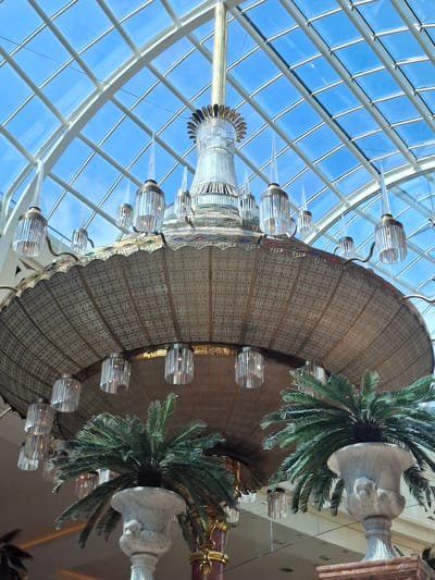 This image captures a low-angle view of a grand, ornate chandelier suspended from a glass-panelled ceiling in a modern indoor space. The chandelier is predominantly made of brass or a similar metallic material, featuring intricate patterned metalwork on its large, conical shade. It is adorned with multiple crystal or glass pendant lights that hang downwards. The ceiling is a grid of white metal struts supporting large glass panels, through which a bright blue sky with sparse white clouds is visible.
In the foreground, to the right and left of the chandelier, are large decorative planters. Each planter contains a lush green cycad or palm-like plant. The planters themselves are elaborate, appearing to be made of stone or a similar material, with sculpted decorative elements. The overall impression is one of opulence and grandeur, suggesting a high-end indoor environment such as a shopping centre, hotel lobby, or atrium.
Based on the description of the scene, this location is likely inside the Trafford Centre in Trafford Park, Trafford, Greater Manchester, United Kingdom. The Trafford Centre is renowned for its elaborate architecture, large atrium, and numerous decorative elements, including grand chandeliers and indoor landscaping. The glass ceiling, in particular, is a prominent feature of the shopping centre.