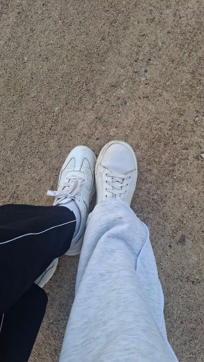 The image shows a close-up view of two pairs of white sneakers and the lower legs of the wearers, standing on a sandy or gravelly surface. One person is wearing black sweatpants with a white stripe down the side and a white athletic shoe. The other person is wearing light gray sweatpants and a white casual sneaker. The surface beneath their feet is a coarse-grained, light brown material, consistent with a pathway or unpaved area. The overall impression is casual and outdoors. The location is identified as Stowbtsy, Belarus.