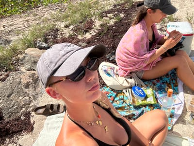 The image depicts two females engaged in leisure activities on a sunny beach in Le Moule, France.
In the foreground, a female is positioned facing the camera, wearing a grey baseball cap, dark sunglasses, a black bikini top, and a layered gold necklace featuring multiple charms including a shell and a mermaid or fish tail. Her blonde hair is partially visible. She is seated on a light green towel. To her left, a single brown Birkenstock-style sandal, labeled "CTWK," rests on the sand.
In the mid-ground, to the right and slightly behind the foreground individual, a second female is seated. She wears a dark grey baseball cap, a pink patterned wrap over a black bikini top, and light-colored shorts. She is looking downwards, actively handling a black camera or small electronic device. A white soft cooler with red trim is situated to her right.
Between the two individuals, on a blue and orange patterned beach towel, are various items: an oval blue tin labeled "DELICIEUX," a green bag of snacks, a clear glass bottle with an orange and green label, and two clear plastic food containers.
The environment consists of a sandy beach surface interspersed with light-colored rocks, patches of green vegetation, and significant accumulations of dark red-brown seaweed.