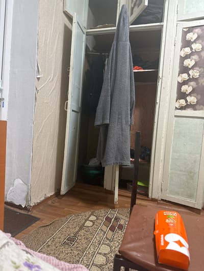 The image depicts an interior scene, likely a room in a residential building, possibly located in Corlăteni, Moldova. The primary focus is a partially open, old-fashioned wooden wardrobe with a gray robe hanging inside. The wardrobe doors are painted a light, faded color, and one door is ajar, revealing shelves within. One of the wardrobe doors has a pane of frosted glass with a floral pattern. To the left of the wardrobe, a wall is visible, covered in peeling wallpaper with a beige, textured pattern. The floor is wooden, with a patterned rug laid down. In the foreground, a brown stool or chair is partially visible, with an orange food package on it. The overall impression is one of a slightly cluttered and worn interior. There are no people visible in the image.