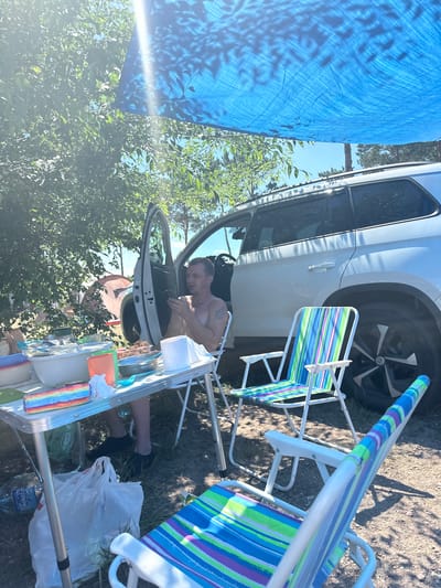 The image depicts a man sitting in a beach chair next to an open car door, looking at his phone. A foldable table is set up in front of him, laden with food containers, bowls, cups, and what appears to be a loaf of bread and a rainbow-colored object. Behind the table, a white SUV is parked, with its rear door also open. The scene suggests a picnic or camping outing. Two colorful striped beach chairs are in the foreground. The environment appears to be outdoors, with trees and foliage visible, and a blue tarp stretched overhead, casting dappled shadows. The ground is covered in dirt and dry grass. The overall impression is a casual, outdoor gathering. No specific details indicate the location in the city of Жемчужный, Russia, beyond it being an outdoor recreational setting.