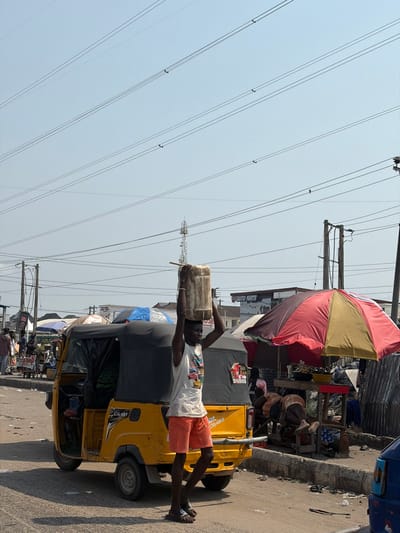 The scene depicts an outdoor street or market area in Ikota, Nigeria, under a clear, light blue sky. Numerous parallel power lines extend horizontally across the upper portion of the frame. In the foreground, a dark-skinned male, wearing a white graphic tank top, orange shorts, and dark flip-flops, walks rightward. He carries a light-colored, cylindrical container on his head, steadying it with both hands.
Behind the male subject, a yellow three-wheeled motorized rickshaw (Keke NAPEP) with a black soft top and items, including blue tarpaulin, stacked on its roof is parked. To the right of the frame, a large red and cream-striped umbrella provides shade over a ground-level market stall displaying various items, including produce. A structure made of corrugated metal sheeting is visible directly behind this stall.
In the mid-ground and background, other individuals are visible, with at least one person appearing to be resting beneath the market umbrella. Various utility poles are present, and a tall, lattice-style antenna tower is visible in the mid-background. A sign bearing the text "AUTO PARTS" is displayed on a building further in the background. The ground consists of an uneven, unpaved or dirt-covered surface.