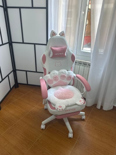 An indoor shot shows a pink and white gaming chair with cat ear accents. The chair is positioned on a wooden floor in what appears to be a room with a window and white sheer curtains. A white folding screen with black geometric dividers is to the left of the chair. The chair has a white faux fur seat cushion with pink paw print accents and a matching back pillow shaped like a cat's paw. The chair is branded "MEENICE". The window shows a brick building outside, and a radiator is visible beneath the window. The setting is likely a domestic interior in Oviedo/Uviéu, Spain.