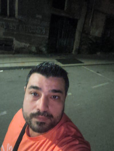 A man with dark hair and a beard, wearing an orange shirt, takes a selfie outdoors at night. He is positioned in the foreground, with a street and building in the background. The building appears to have graffiti on its wall. The street is paved and has visible white lines. The lighting suggests an urban environment. It is not possible to determine the exact location within Harmanli, Bulgaria from the provided image.