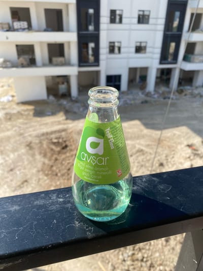 A glass bottle of Avşar brand "Yeşil Elma" (Green Apple) flavored sparkling mineral water lies on its side, leaning against a dark blue, possibly metal surface. The bottle has a green label with white text. The visible text on the label includes "Yeşil Elma" and "avsar", "Yeşil elma aromalı doğal zengin mineralli gazlı içecek", and "Afyonkarahisar". There is also text that appears to be nutritional information and ingredient details. The liquid inside the bottle is clear and appears to have carbonation.
In the background, a white building with multiple floors and dark-framed windows is visible, appearing to be under construction or renovation. The building's facade is stark and modern. The ground appears to be dirt or rubble, consistent with a construction site. The lighting suggests it is daytime, with shadows cast on the ground. The overall scene suggests a casual setting, perhaps outdoors at a construction site or a balcony overlooking one. There are no people visible in the image.