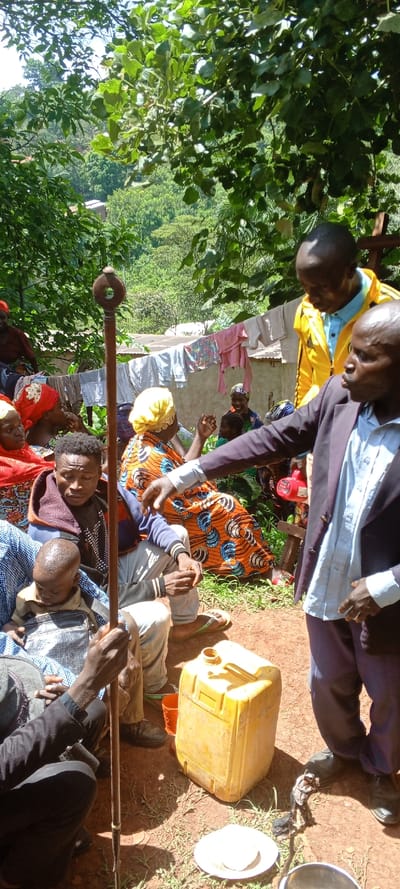 A group of people are gathered outdoors. Some sit, others stand, near a yellow container and a plate. A long, thin staff with a circular top is held by a person in the foreground. Clothes hang on a line in the background near a building. Some of the people are dressed in colorful patterned fabrics, while others wear jackets and trousers. A man in a yellow jacket speaks to another man in a blue shirt and a blazer.