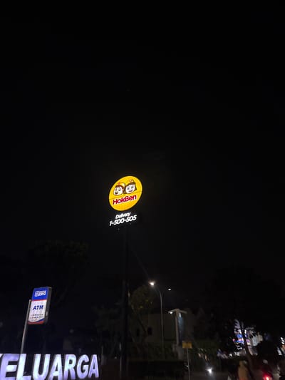 This media file depicts a nighttime scene in South Tangerang, Indonesia. The primary subjects are illuminated signs. A prominent, circular yellow sign features the "HokBen" logo, which includes two cartoon characters and the text "HokBen" and "Delivery 1-500-505". To the right of this, a large, white, illuminated sign displays the word "KELUARGA" in capital letters. Below the HokBen sign, a smaller blue and white sign indicates "BRI ATM". The setting appears to be a street or commercial area at night, with streetlights visible in the background and the ambient darkness suggesting it is after sunset. There are no people clearly visible. The overall mood is that of an urban commercial landscape at night, with the bright lights of the signs contrasting with the dark surroundings.
