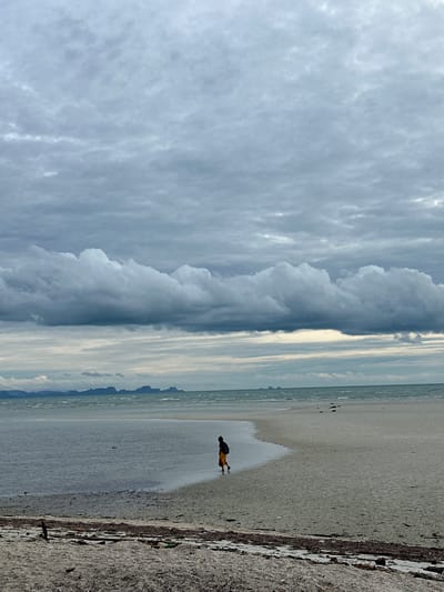 The image shows a wide, flat, sandy beach under a heavily overcast sky. A person in a dark top and yellow skirt is walking away from the camera along the water's edge, with their back to the viewer. The water is calm and shallow, and the horizon is marked by a line of distant islands or peninsulas with jagged peaks. The foreground of the beach is strewn with debris, including seaweed and small pieces of wood. The overall mood is somber and serene. This scene is consistent with a quiet beach on Ko Samui District, Thailand, likely during the monsoon season.
