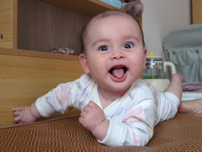 A baby is lying on its stomach on a textured brown surface. The baby is wearing a white onesie with a floral pattern. Its mouth is open and tongue is visible. The baby's eyes are wide. In the background, there is a light-colored wooden cabinet or shelf, and other indistinct objects are out of focus. The image does not contain identifiable landmarks to confirm its specific location in Shunde District, China.