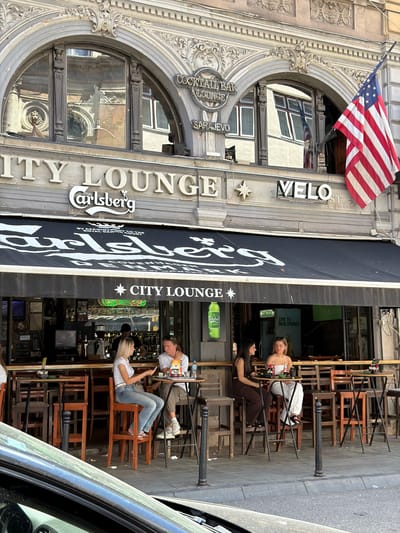 The image shows the exterior of a bar or lounge named "City Lounge" located in Sarajevo, Bosnia and Herzegovina. The establishment features a prominent black awning with the "Carlsberg" logo printed in white, along with the text "BY APPOINTMENT TO THE ROYAL DANISH COURT COPENHAGEN DENMARK". Above the awning, the words "CITY LOUNGE" are displayed in large, light-colored lettering, accompanied by a star symbol and the word "VELO" to its right. An American flag is visible hanging from the building on the right side of the frame.
In front of the establishment, several people are seated at tables and high chairs on the sidewalk. On the left, two women are seated at a high table. One woman, with blonde hair, is wearing a white top and jeans, and is looking at her phone. The other woman, with dark hair, is wearing a dark top and brown pants. Further to the right, a man and a woman are seated at a round table. The man has blonde hair and is wearing a white t-shirt and khaki pants, and is looking at the woman. The woman is wearing a white strapless top and white pants.
The background reveals a street scene with other buildings and windows reflected in the upper windows of the "City Lounge". There are bar shelves visible inside, stocked with bottles and glasses, and a person is working behind the bar. A green bottle of Carlsberg beer and a bottle of Somersby are also visible outside the bar. The overall impression is a lively outdoor seating area of a bar in an urban setting.