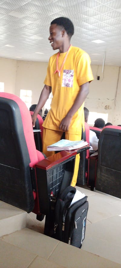 In a room with a tiled ceiling and cream-colored walls, a young man wearing a yellow traditional outfit stands in front of a row of red and black chairs, typical of an auditorium or lecture hall. He has a bright smile on his face and is wearing a pink necklace with a decorative pendant. His outfit appears to be a tunic and pants set. On the armrest of a chair in the foreground, a stack of papers or books is placed. A black backpack with a white accent is visible on the floor next to the chair. The room is dimly lit, with light filtering in from windows in the background, suggesting it is daytime. The overall atmosphere appears casual, possibly a lecture or gathering within an educational institution. The city of Gumel, Nigeria, is characterized by its warm climate and educational institutions, making this scene plausible within that context.