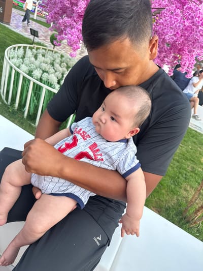 A man is holding a baby, seen from the chest down. The man is wearing a black t-shirt and dark pants with a "Demix" logo. The baby is wearing a grey and white striped onesie with red lettering that appears to say "BASE BALL" and the number "3". The baby is looking down and to the right, with a neutral expression. The man is looking down at the baby with his eyes closed, appearing to be nuzzling the baby.
In the background, there are decorative pink flowers on a tree to the left. To the right, there are white, fluffy decorative plants and green grass. The scene suggests an outdoor setting, possibly a park or garden. The lighting indicates it is daytime. The overall mood is one of tenderness and affection between the man and the baby. The location is identified as Manas, Kyrgyzstan.