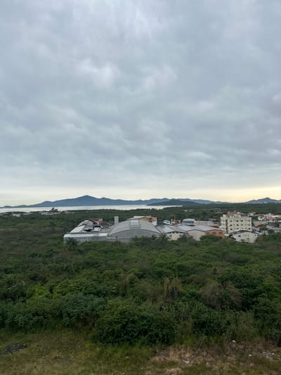 The image is a wide-angle, landscape-oriented photograph taken from an elevated position, looking down on a coastal town. The scene is divided by a horizon line: the left two-thirds are dominated by a cloudy, overcast sky, while the right third depicts a lush, green hillside dotted with buildings and overlooking a body of water and distant mountains.

The hillside is densely covered in trees and vegetation, with various structures interspersed. Closer to the viewer, there's a modern building with a curved, white roof, possibly a stadium or a large hall. Further up the hill, more buildings are visible, some appearing to be apartment complexes or hotels with multiple stories and visible windows. The architecture suggests a tropical or subtropical climate.

Below the hillside, a calm body of water stretches towards the horizon, flanked by a series of progressively larger, darker mountain ranges. The water reflects the muted light of the sky, creating a serene and slightly hazy atmosphere. The time of day appears to be late afternoon or early morning, given the soft, diffused light and the overcast conditions. The weather is clearly cloudy and somewhat dull.

The overall impression is of a developing coastal community nestled between verdant hills and the sea. There is no visible text in the image. The scene evokes a sense of tranquility and natural beauty, juxtaposed with human development.