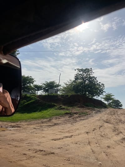 The image captures a view from a vehicle, likely a tuk-tuk, looking out at a dirt road in Karmajiji, Nigeria. The road is rutted and appears to be somewhat muddy in places, indicating recent rainfall. The left side of the frame shows a reflection in a side mirror, revealing a person's shoulder and arm, possibly the driver or passenger.
The environment is lush with green grass and several trees dotting the landscape. Some structures, possibly small buildings or utility boxes, are visible behind the trees on a raised embankment. The sky is bright with scattered white clouds and the sun is high, casting a glare. The overall impression is of a rural or semi-urban setting on a sunny day.