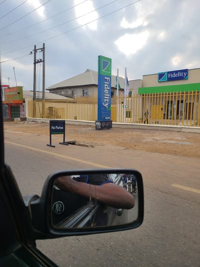 The media file depicts a street scene in Karu, Nigeria. The primary focus is a building with signage for "Fidelity," a financial institution, with its green and blue logo visible. A yellow metal fence surrounds the property. In the foreground, a side-view mirror of a vehicle reflects a person's arm and a motorcycle. The road surface appears to be asphalt with yellow markings. A "No Parking" sign is also visible. Power lines and poles indicate urban infrastructure. The sky is cloudy, suggesting a potentially overcast day. The overall impression is that of a typical street view in a populated area, possibly during daylight hours given the brightness. The scene appears to be dynamic, with the implication of a vehicle in motion.