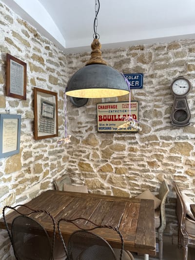 The image depicts an indoor scene with a rustic ambiance, possibly a cafe or restaurant, with a prominent stone wall. A wooden table with chairs is centered in the foreground. Above the table, a large industrial-style pendant lamp hangs, casting a warm glow. The stone wall is adorned with several framed items and signs, including what appears to be a vintage clock on the right and a sign with French text that reads "CHAUFFAGE PLOMBERIE ENTRETIEN SANITAIRE BOUILLARD." There are also framed documents and a sign for "BISCUITS PERNOT" on the left. Two chairs with woven seats and metal frames are positioned at the table. The overall environment suggests a cozy and historical setting. There are no people visible in the image. The location in Semogorskiy, Russia is not discernible from the visual cues provided in the image.