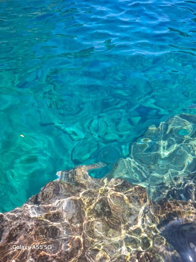 The image shows a view of the clear, turquoise water of the sea near Plakias, Greece. A rocky outcrop is visible on the right side of the frame, partially submerged, with light reflecting off its surface. The water's surface is textured with ripples and waves, creating varied patterns of light and shadow. A small, yellowish object floats in the water. The scene is naturally lit with sunlight. The bottom right of the image is marked with "Galaxy A55 5G."