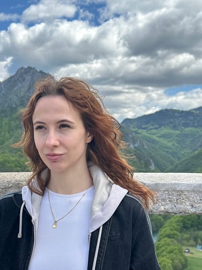 A reddish-haired woman in a white t-shirt and black jacket stands beside a stone railing. Behind her are mountains and a cloudy sky. The location is Đurđevića Tara, Montenegro.