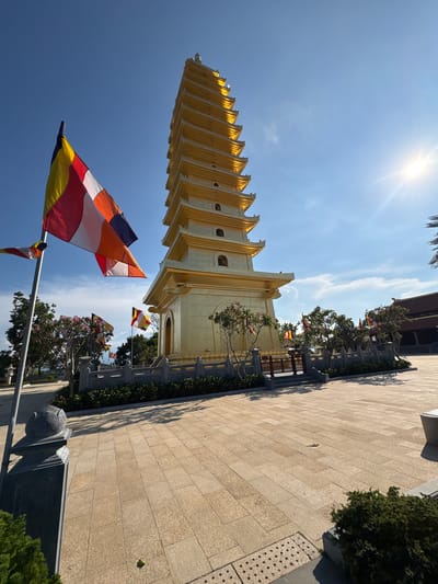 The image shows a tall, golden pagoda located in Diên Khánh, Vietnam. The pagoda has multiple tiers and a distinctive architectural design. In the foreground, several Buddhist flags with horizontal stripes in red, yellow, white, orange and blue are waving in the wind. The sky is bright blue with a few clouds and the sun shining at the upper center. To the side of the pagoda, there is a paved walkway lined with trees and shrubs.