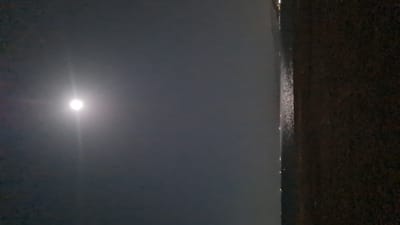 This nighttime image, taken in Eilat, Israel, captures a serene moonlit scene over a body of water. The photograph is oriented vertically, with the primary light source, a brilliant white moon, positioned towards the upper-left quadrant. The moon is exceptionally bright, producing a strong, vertical lens flare or glow that extends downwards across the dark sky. Its clarity indicates a clear night.
To the right of the moon, the dark expanse of the sea or ocean fills the majority of the frame. This water surface appears calm and subtly reflective, with faint glimmers of light scattered across it, likely reflections from the moon or distant sources.
On the far right, a textured, dark vertical mass occupies approximately a quarter of the image. This could be a rocky coastline, a concrete promenade, or the edge of a structure overlooking the water. Along the interface between this dark mass and the water, a scattering of small, distinct lights is visible, suggesting distant city lights, coastal infrastructure, or lights from boats.
The overall scene is tranquil and devoid of human activity, focusing instead on the natural beauty of the moonlit night and the vastness of the sea, subtly juxtaposed with the distant lights of civilization. No visible text is present in the image.