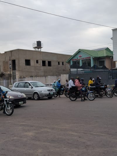 A street scene in Kabala, Nigeria. Multiple motorcycles are parked along the road, some with riders. Several cars are also parked near the motorcycles. Buildings of varying architectural styles are visible in the background. The sky is overcast.